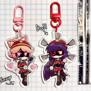 以下が含まれることがあります： ピンクと赤の留め具が付いた、漫画のキャラクターのアクリルキーホルダー2点。左のキャラクターは明るい髪でピンクの留め具、Lizzyと表記。右のキャラクターは紫の髪で赤の留め具、Dollと表記。定規は高さ10cmを示している。