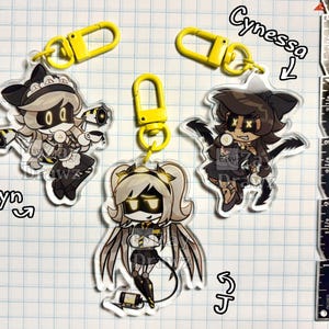 以下が含まれることがあります： Cyn、Cynessa、Jとラベル付けされた漫画キャラクターの透明アクリルキーホルダー3個。それぞれに黄色のプラスチック製ロブスタークラスプが付いています。右側の金属製定規はインチ単位の目盛りを示しており、キーホルダーの高さは約6.35cmです。