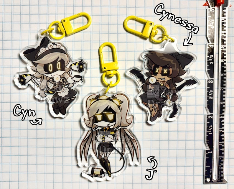 以下が含まれることがあります： Cyn、Cynessa、Jとラベル付けされた漫画キャラクターの透明アクリルキーホルダー3個。それぞれに黄色のプラスチック製ロブスタークラスプが付いています。右側の金属製定規はインチ単位の目盛りを示しており、キーホルダーの高さは約6.35cmです。