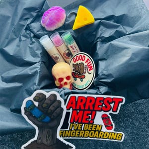 May include: A collection of novelty items including a pink brain-shaped item, a yellow cheese wedge, lip balms, a skull, and stickers. The stickers feature a hand holding a skateboard and the text "ARREST ME! I'VE BEEN FINGERBOARDING."