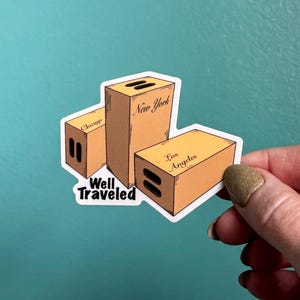 May include: A sticker featuring a stack of three cardboard boxes labelled "Chicago", "New York", and "Los Angeles". The boxes are stacked on top of each other and the sticker says "Well Traveled" below the boxes.