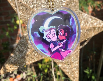 May include: A heart-shaped acrylic charm with a purple, blue, and pink illustration of two cartoon characters holding hands under a crescent moon. The charm is on a gold chain.