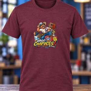 May include: A maroon short-sleeved t-shirt featuring a graphic of a cartoon worker with a mustache and straw hat giving a thumbs up. The design includes tools, a toolbox, and the text HAGO CHAPUCES! in bold yellow and white lettering.