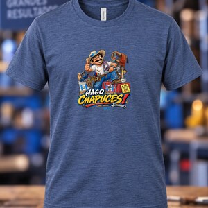 May include: A heather blue short-sleeve t-shirt featuring a graphic of a cartoon worker with a mustache and hat, surrounded by tools and a red toolbox. The text HAGO CHAPUCES is printed in bold yellow and white letters below the illustration.