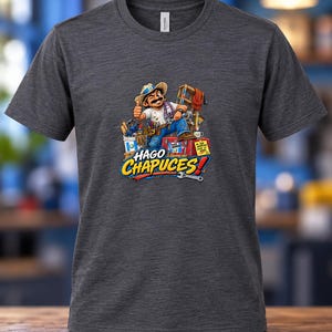 May include: A dark heather grey short-sleeve t-shirt featuring a cartoon illustration of a construction worker giving a thumbs up, surrounded by tools and a ladder. The text HAGO CHAPUCES is printed in large, bold, colorful letters below the character.