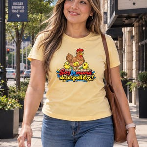 May include: A light yellow short-sleeve t-shirt featuring a graphic of a brown hen with three yellow chicks and the text Soy la mama de los pollitos in colorful lettering. The shirt is paired with blue denim jeans and a brown shoulder bag.