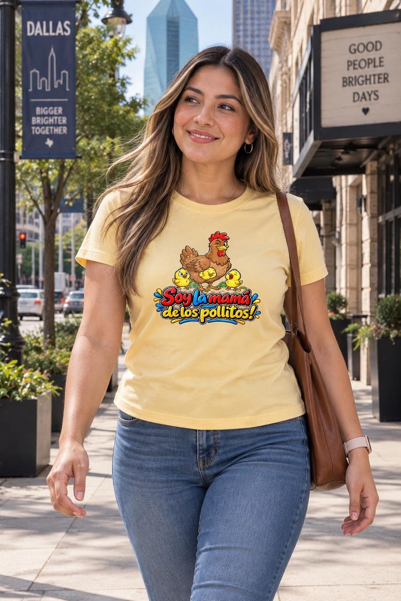 May include: A light yellow short-sleeve t-shirt featuring a graphic of a brown hen with three yellow chicks and the text Soy la mama de los pollitos in colorful lettering. The shirt is paired with blue denim jeans and a brown shoulder bag.