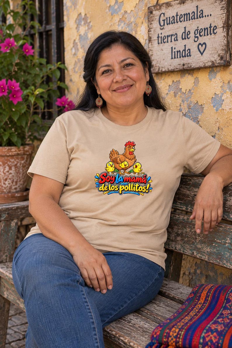 May include: A beige short-sleeved t-shirt featuring a graphic of a brown hen with three yellow chicks. Below the graphic, the text reads "Soy la mam&aacute; de los pollitos!" in colorful, stylized lettering with blue, red, and yellow accents.