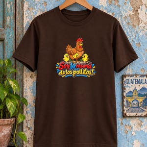May include: A dark brown short-sleeve t-shirt featuring a graphic of a brown hen with three yellow chicks. Below the illustration, the text reads "Soy la mam&aacute; de los pollitos!" in colorful, stylized lettering with blue, red, and yellow accents.