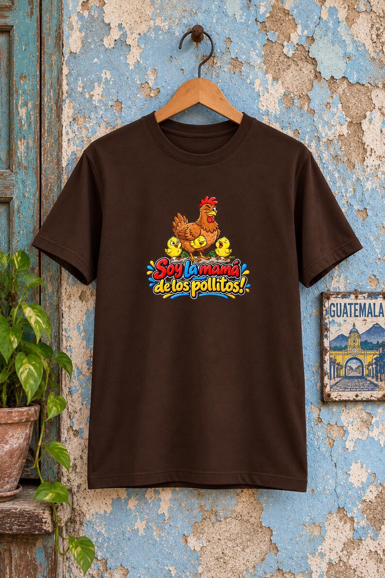 May include: A dark brown short-sleeve t-shirt featuring a graphic of a brown hen with three yellow chicks. Below the illustration, the text reads "Soy la mam&aacute; de los pollitos!" in colorful, stylized lettering with blue, red, and yellow accents.
