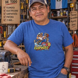 May include: A blue short-sleeved t-shirt featuring a graphic illustration of a cartoon worker with tools and the text HAGO CHAPUCES. The design includes a bucket, a ladder, and a red toolbox. The shirt has a crew neckline and a heathered texture.