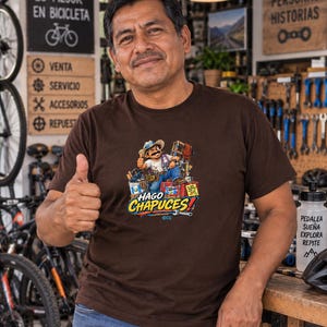 May include: A brown short-sleeved t-shirt featuring a graphic illustration of a cartoon character wearing a hat and blue jeans, surrounded by tools and red crates, with the text "HAGO CHAPUCES!" printed in large yellow and blue letters.
