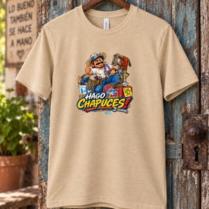 May include: A tan short-sleeve t-shirt featuring a graphic of a cartoon construction worker giving a thumbs up, surrounded by tools and a ladder. The text HAGO CHAPUCES! is printed in large blue and yellow letters below the illustration.