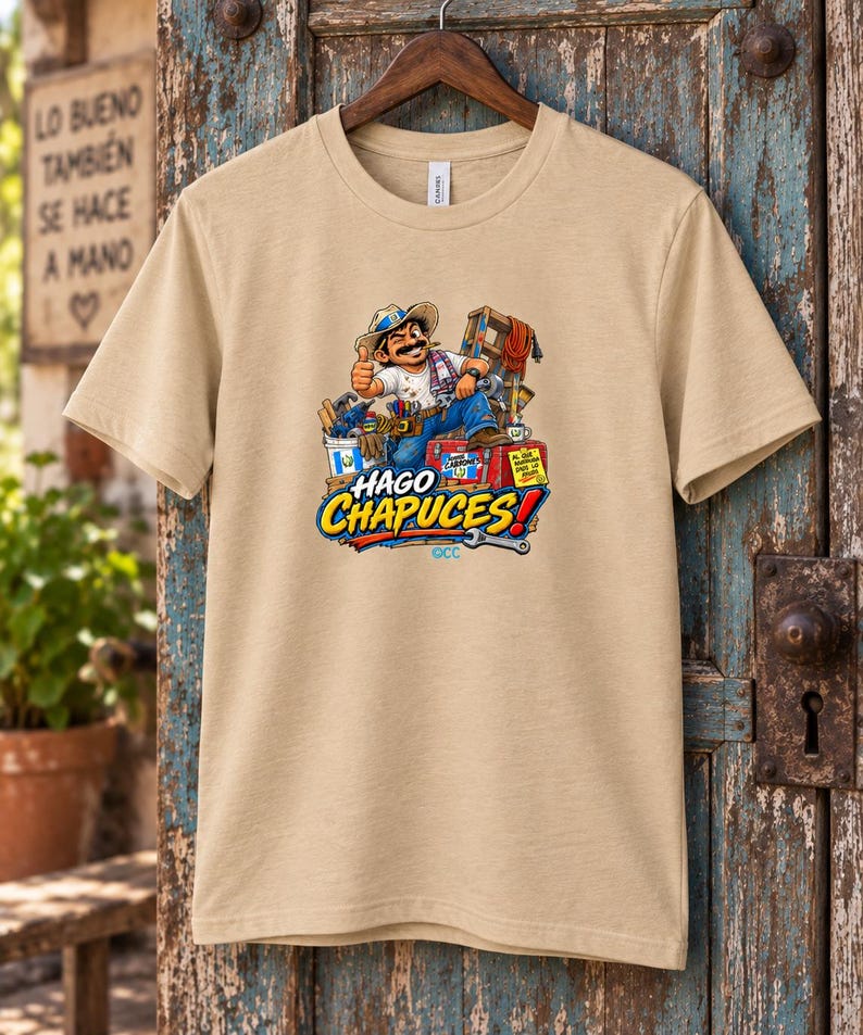 May include: A tan short-sleeve t-shirt featuring a graphic of a cartoon construction worker giving a thumbs up, surrounded by tools and a ladder. The text HAGO CHAPUCES! is printed in large blue and yellow letters below the illustration.