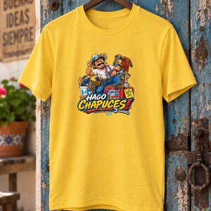 May include: A bright yellow short-sleeved t-shirt featuring a graphic illustration of a cartoon worker with a mustache and hat, surrounded by various tools and a red toolbox. The text HAGO CHAPUCES is printed in large blue and yellow letters below.