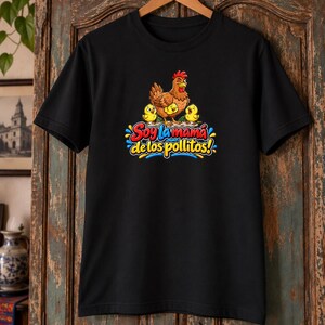 May include: A black short-sleeved t-shirt featuring a graphic of a brown hen with three yellow chicks. Below the illustration, the text reads "Soy la mam&aacute; de los pollitos!" in colorful, bold, stylized lettering with blue and yellow accents.