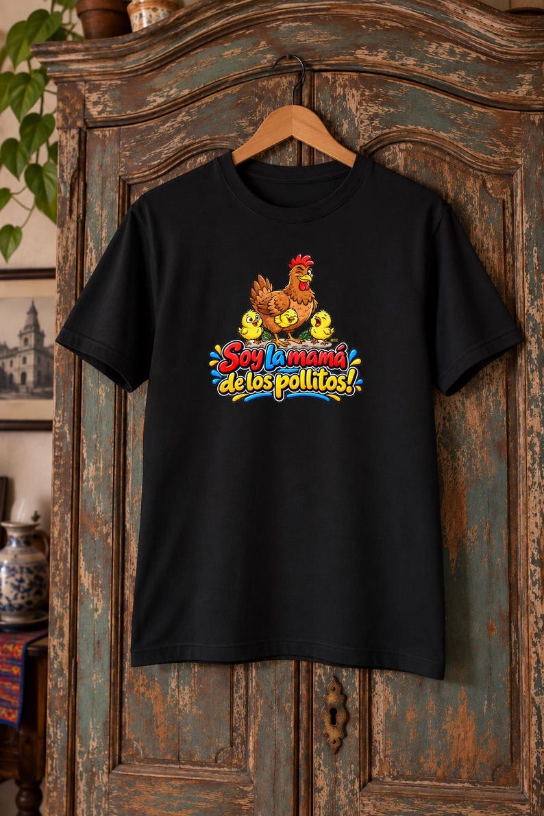 May include: A black short-sleeved t-shirt featuring a graphic of a brown hen with three yellow chicks. Below the illustration, the text reads "Soy la mam&aacute; de los pollitos!" in colorful, bold, stylized lettering with blue and yellow accents.