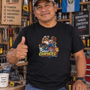 May include: A black t-shirt featuring a colorful graphic of a cartoon character wearing a hat and tool belt, surrounded by tools and a toolbox, with the text "HAGO CHAPUCES!" printed in bold yellow and blue letters below the illustration.