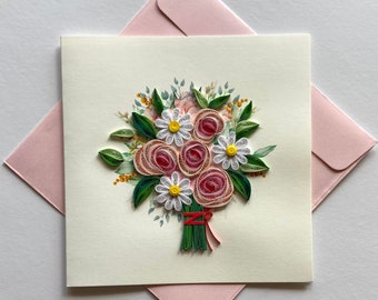 May include: A handmade greeting card featuring a quilled paper bouquet of pink roses and white daisies with yellow centers. The card is on a cream background, with a pink envelope behind it. The bouquet is tied with a red ribbon.