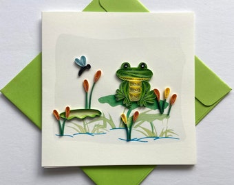 May include: A white card featuring a green frog sitting on a lily pad. The frog is crafted from paper and has yellow accents. A blue dragonfly is flying above the frog.