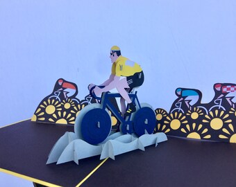 May include: A pop-up card featuring a cyclist in a yellow jersey riding a blue bicycle. The cyclist is surrounded by a field of yellow flowers. The card is made of paper and has a 3D effect.
