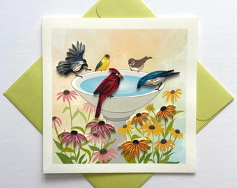 May include: A greeting card featuring a colorful paper craft scene of birds perched on a bird bath surrounded by flowers. The birds are a cardinal, a blue jay, a yellow bird, and a sparrow. The card has a white background with a light brown border.