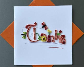 May include: A white greeting card with the word "Thanks" in a decorative, quilled style. The letters are a mix of red, orange, and purple, with floral and insect accents. An orange envelope is visible.
