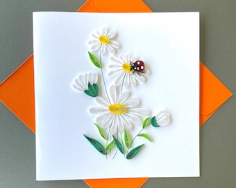 May include: A white card with a quilled paper daisy flower arrangement. The flowers are white with yellow centers and green leaves. A red ladybug is perched on one of the flowers.