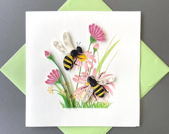 May include: A handmade greeting card featuring two paper quilled bees hovering over pink flowers and green grass. The card is on a white background, with a light green envelope in the background. The bees have black and yellow stripes.
