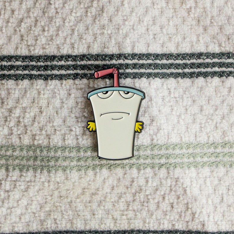 May include: A white enamel pin of a cartoon character, a milk carton with a straw, with a blue lid and yellow arms. The character has a sad expression and is wearing a red straw.