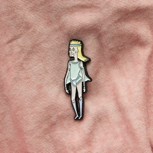 May include: A black and white enamel pin featuring a cartoon character with blonde hair, a white leotard, and a headband. The character has a serious expression and star-shaped eyes.
