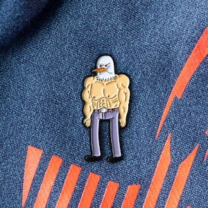 May include: A white eagle head with red eyes and a yellow beak on a muscular, buff, human body wearing blue jeans. The figure is a cartoon-style enamel pin.