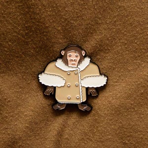 May include: A pin depicting a cartoon ape in a tan coat with white fur trim and a double-breasted design. The ape has a brown face and feet. The pin is set against a brown background.