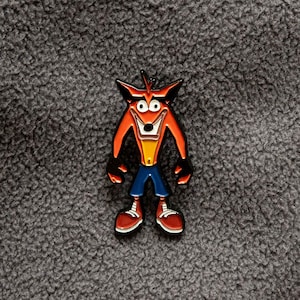 May include: A colorful enamel pin featuring the character Crash Bandicoot. The pin depicts Crash with his signature orange fur, black arms and legs, blue pants, and red shoes. The pin is set against a gray, textured background.
