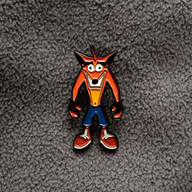 May include: A colorful enamel pin featuring the character Crash Bandicoot. The pin depicts Crash with his signature orange fur, black arms and legs, blue pants, and red shoes. The pin is set against a gray, textured background.