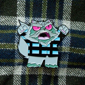 May include: A grey enamel pin of a cartoon cat with a blue and black checked shirt, angry expression, and pink eyes. The cat is wearing a checked shirt and has a white beard.