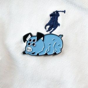 May include: A blue enamel pin of a cartoon pig with five eyes and a black outline. The pig is lying on its belly with its legs outstretched.