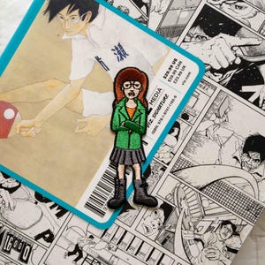 May include: An embroidered patch of a cartoon character with brown hair, wearing a green jacket, a black skirt, and black boots. The character is standing with their arms crossed. The patch is on a book with a black and white manga illustration. The book cover has the text "VIZ SIGNATURE MEDIA" and "ISBN: 978-1-9747-1165-9".