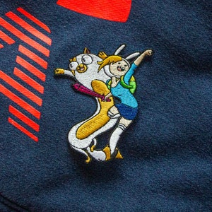 May include: Embroidered patch featuring a cartoon character riding a dog-like creature. The character is wearing a blue shirt and a pink skirt. The dog-like creature is white with a yellow body. The patch is on a dark blue background.