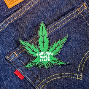 May include: Green and white embroidered patch of a maple leaf with the text "Toronto Maple Leafs" on a blue denim background.