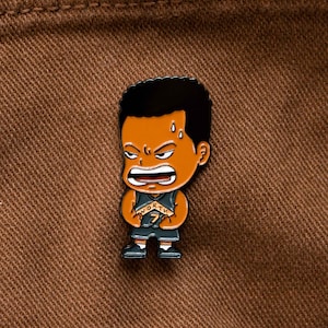 May include: A black and orange enamel pin of a cartoon basketball player wearing a jersey with the word "NORTH" and the number "7" on it. The player has a determined expression on his face and is sweating.