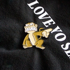 May include: A yellow and black enamel pin featuring a cartoon monkey with a menacing expression and sharp teeth. The monkey is pointing its finger to the right. The pin says "LOVE YO SE" in white text on a black background.