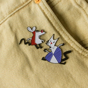 May include: Two enamel pins featuring cartoon animals. The first pin is a white mouse with red and black stripes, and the second pin is a white cat with blue and black stripes.