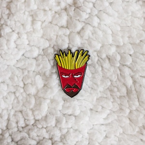 May include: A red and yellow enamel pin featuring a cartoon face with a french fry hairdo. The face has a red skin tone, black eyebrows, and a black mustache and beard.