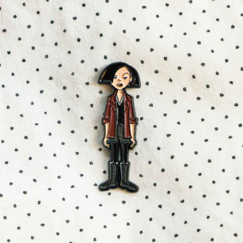 May include: A black and red enamel pin featuring a cartoon character with black hair, a red jacket, and black pants. The character has a serious expression and is wearing black boots.