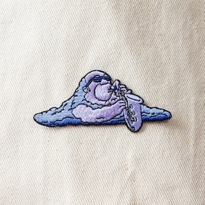 May include: Purple embroidered patch of a cartoon character playing a saxophone. The character is wearing sunglasses and is sitting on a cloud.