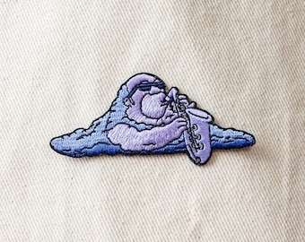 Puede incluir: Parche bordado morado de un personaje de dibujos animados tocando un saxofón. El personaje lleva gafas de sol y está sentado en una nube.