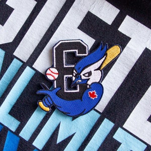 May include: Blue and black embroidered patch of a cartoon blue jay holding a baseball bat with a baseball and the letter "E" in the background. The patch has a red maple leaf on the jay's chest.