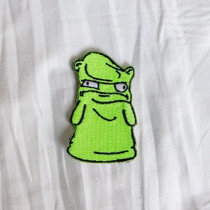 May include: Green embroidered patch of a cartoon character with a grumpy expression. The character has a green body, black eyes, and a black outline.