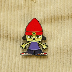 May include: An enamel pin featuring a cartoon dog wearing a red beanie with a green frog logo. The dog has a purple shirt and black trousers.